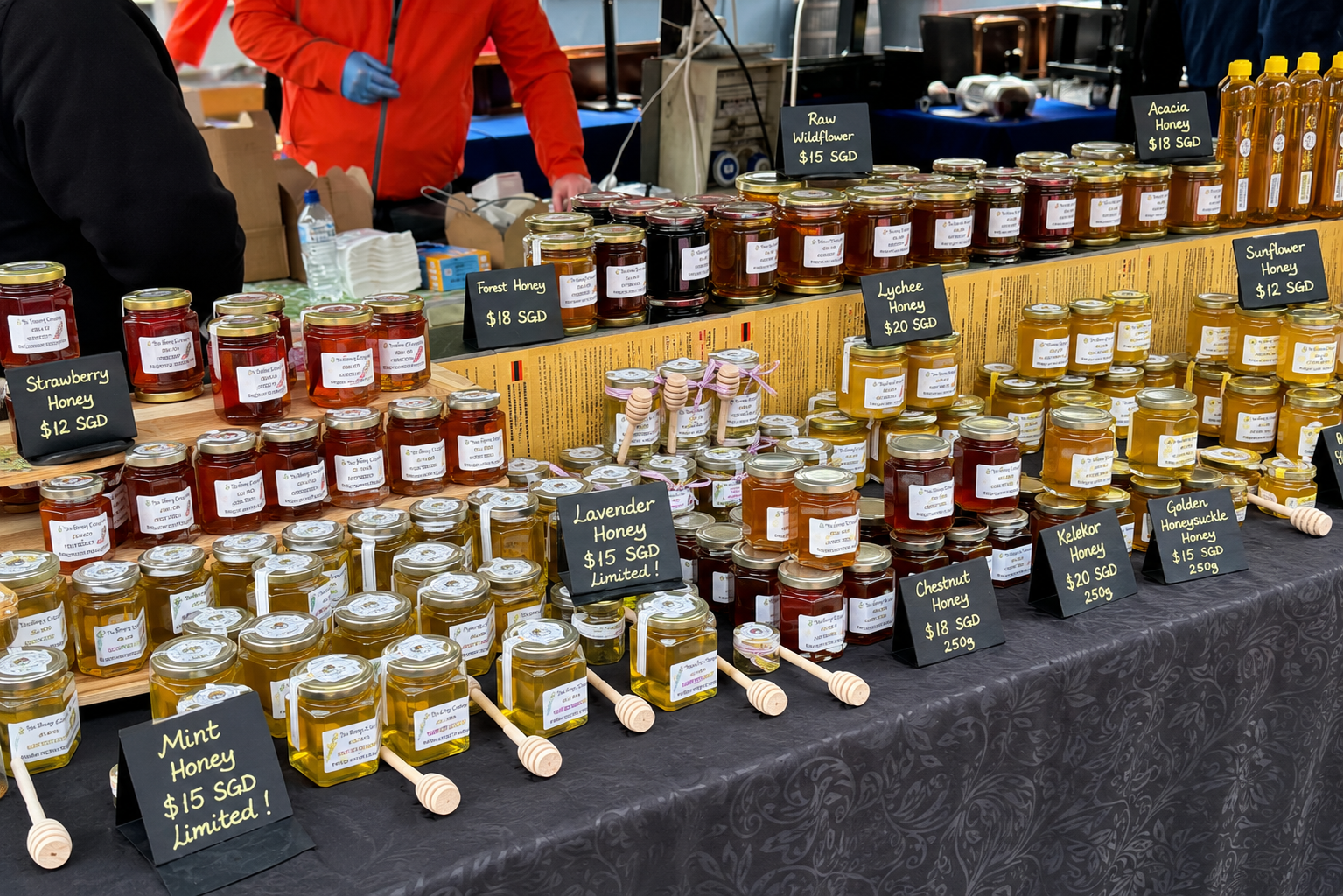 Honey market stall displaying assorted artisanal honey jars with wooden honey dippers and handwritten SGD price signs.