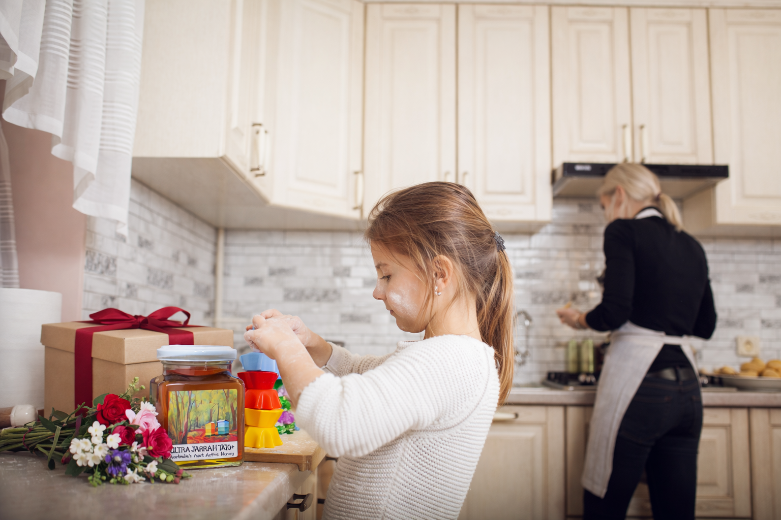 Girl playing in kitchen; gift box, flowers, and honey jar on the counter.