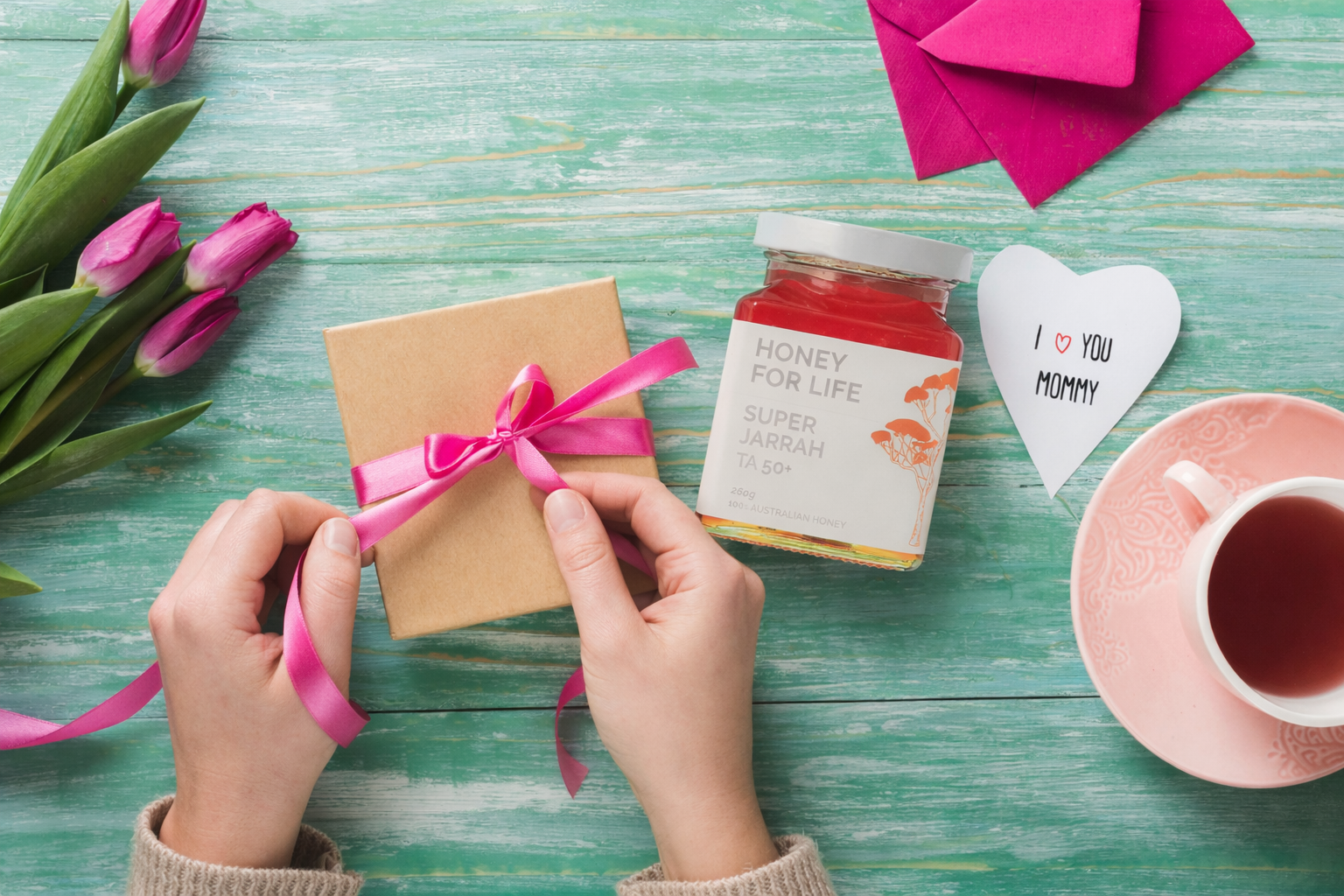 Top-down view of hands tying a pink ribbon around a kraft gift box on a green wooden table, with pink tulips, a heart-shaped “I love you mommy” card, a jar of Honey for Life Super Jarrah honey, and a cup of tea nearby.