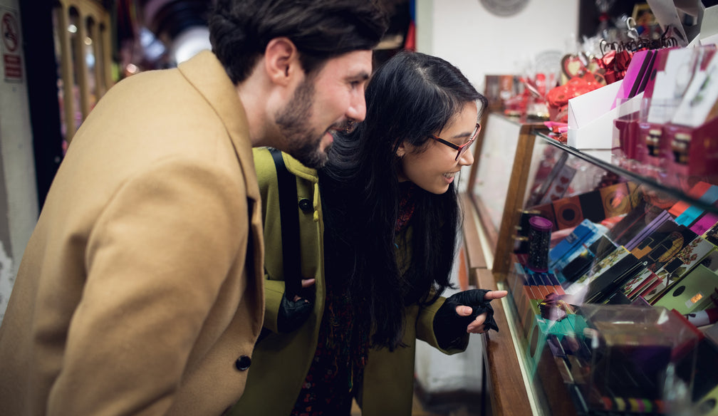 A couple leaning over a glass display case in a gift shop, smiling as they look at colorful boxed items and small packaged gifts inside the counter.