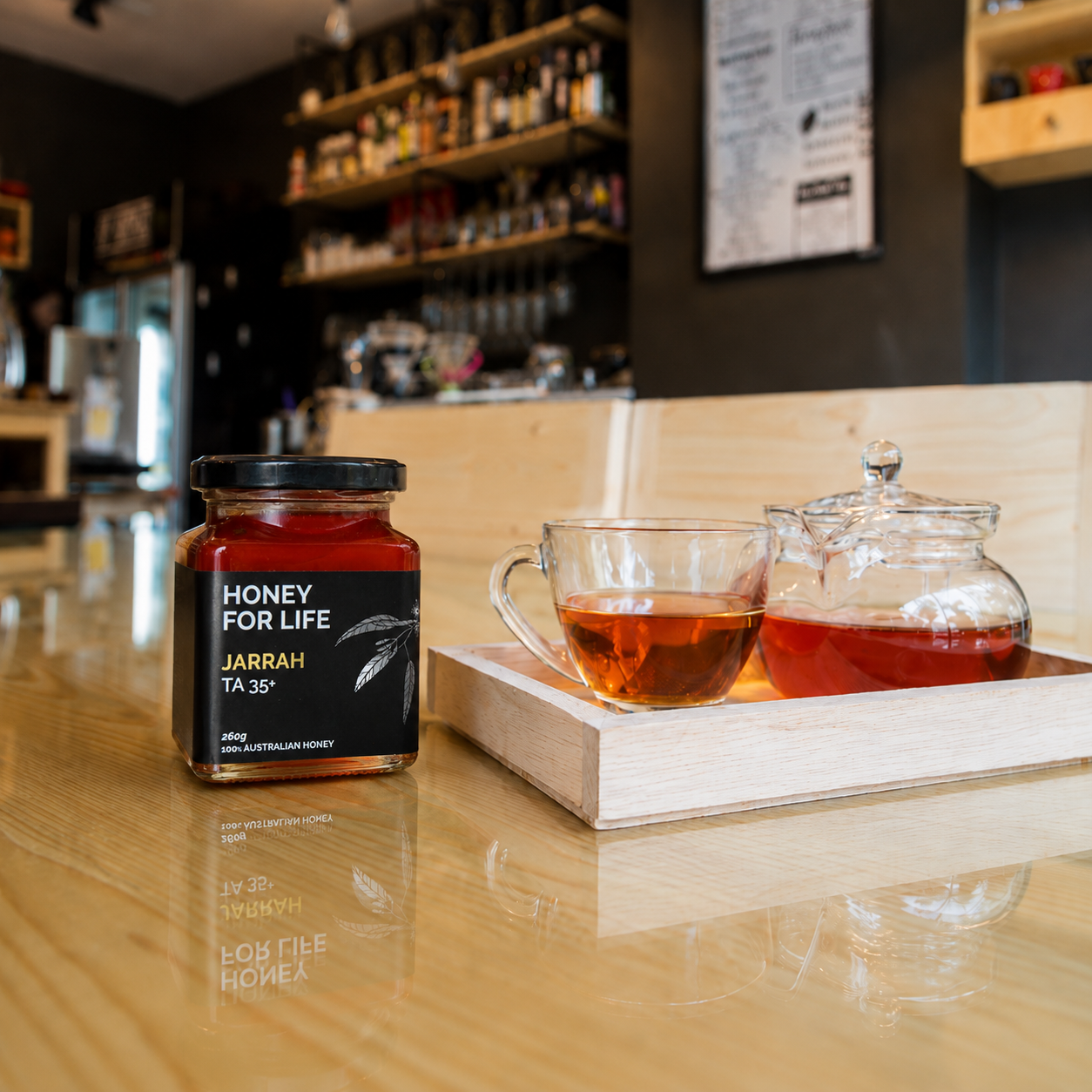 An open luxury black presentation gift box with a gold-lined interior sits on a light wooden table surrounded by eucalyptus leaves, white blossoms, and a glass cup of amber tea.