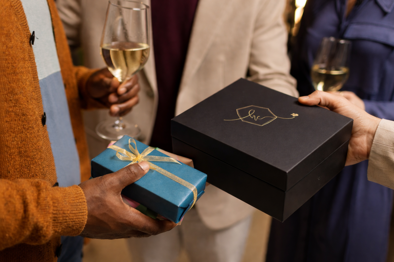 Hands exchanging luxury gift boxes at an elegant celebration, featuring a black Honey Colony gift box and a blue ribbon-wrapped present with wine glasses in the background.