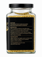 A clear glass jar filled with yellow honey bee pollen granules, featuring a black screw-on lid and a black label with white text and a small gold bee illustration. The back of the label displays product information and a tasting note.